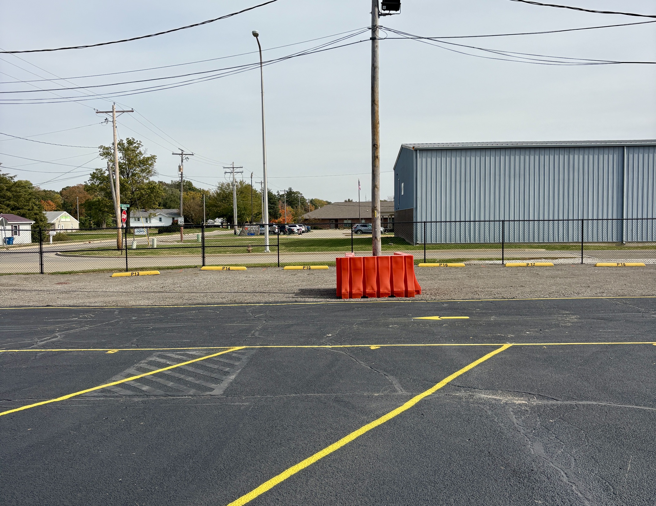Striped paved lot facing outdoor parking spots P13 through P18 along a black chain-link fence beside a metal building