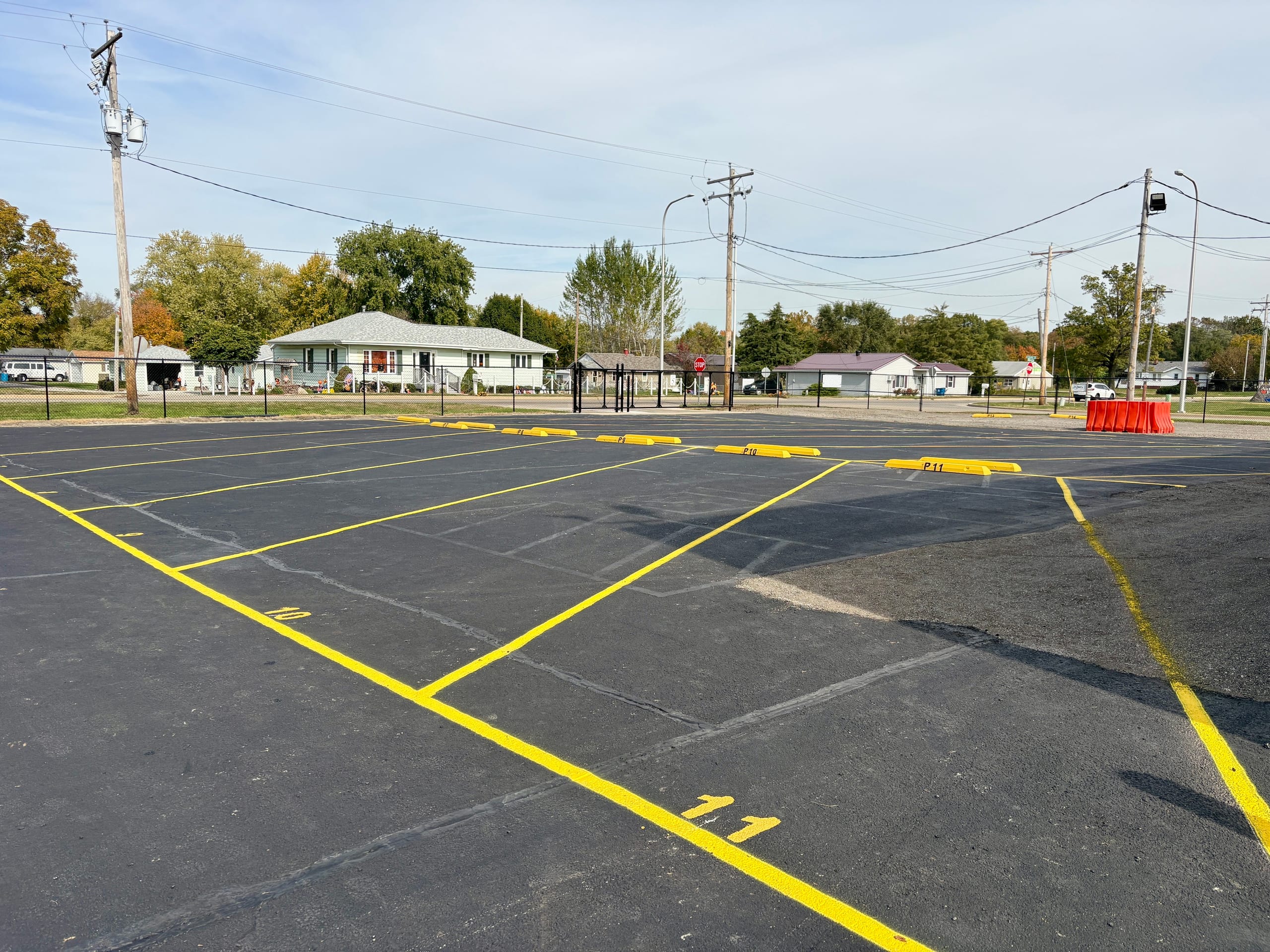 Freshly paved outdoor parking lot with bright yellow striped and numbered spaces, yellow wheel stops, and a black chain-link fence